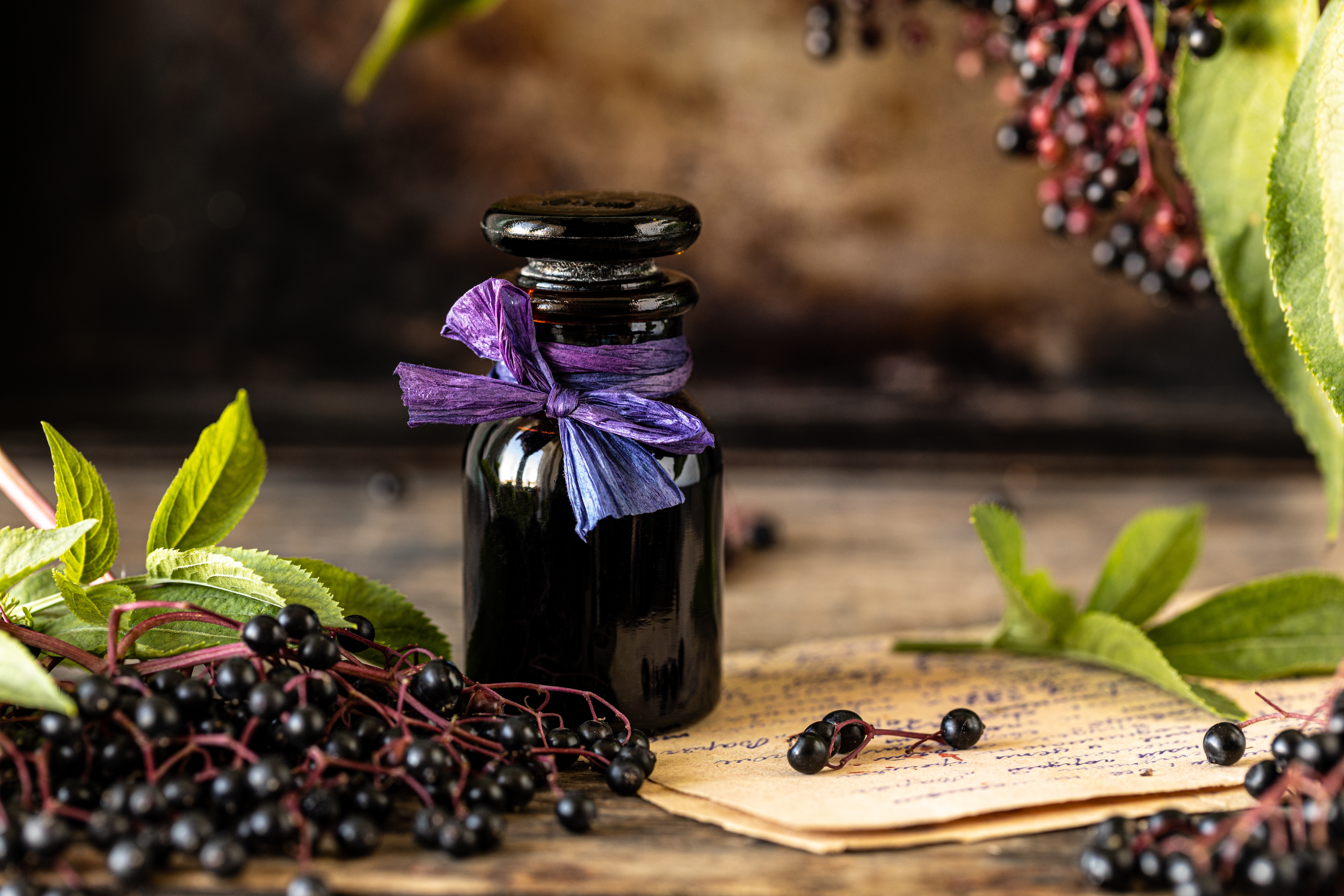 Homemade black elderberry syrup recipe in a glass bottle on a wooden table. Fresh berries in the background